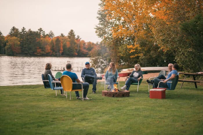 A group of people sit in chairs around a fire pit by a lake on a grassy area with autumn trees in the background.