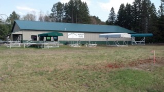 A single-story green-roofed building with canopies, picnic tables, and a sign reading "Camp." Surrounded by trees and grassy area.