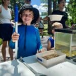 A boy in a blue shirt and hat holds a long net by a table with other children and adults nearby. There are containers with water and a magnifying glass on the table. Trees and sky in the background.