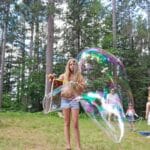 A young girl creates a large soap bubble using a wand in a forested area, with other people in the background.