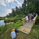 A group of people stands on a wooden bridge overlooking a lush, green landscape with a stream. Two children kneel at the edge of the bridge.