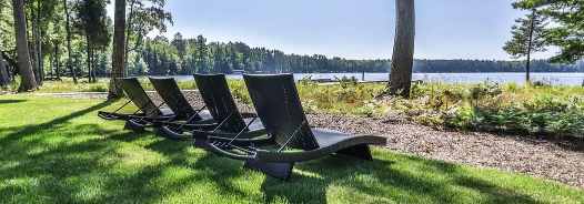 Four black lounge chairs are arranged on a grassy lawn facing a lake, with trees and forested shoreline in the background.
