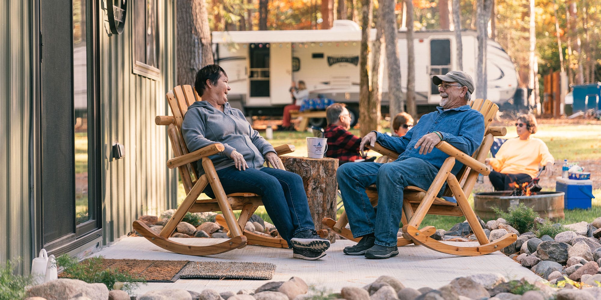 Two people sit on rocking chairs outside a cabin, smiling at each other. A trailer and more people are visible in the background, surrounded by trees.