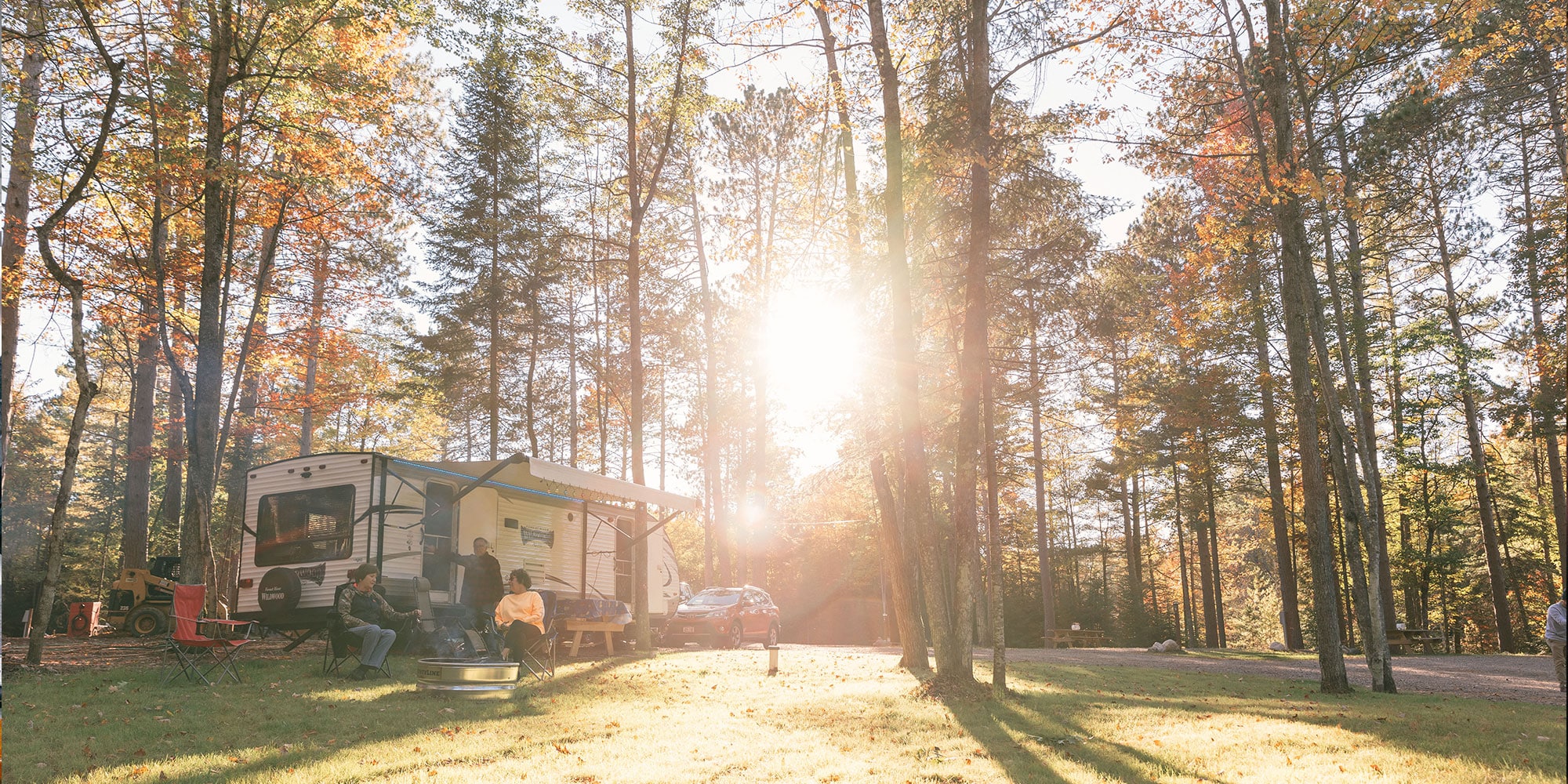 A camper is parked in a forest clearing with sunlight streaming through the trees.