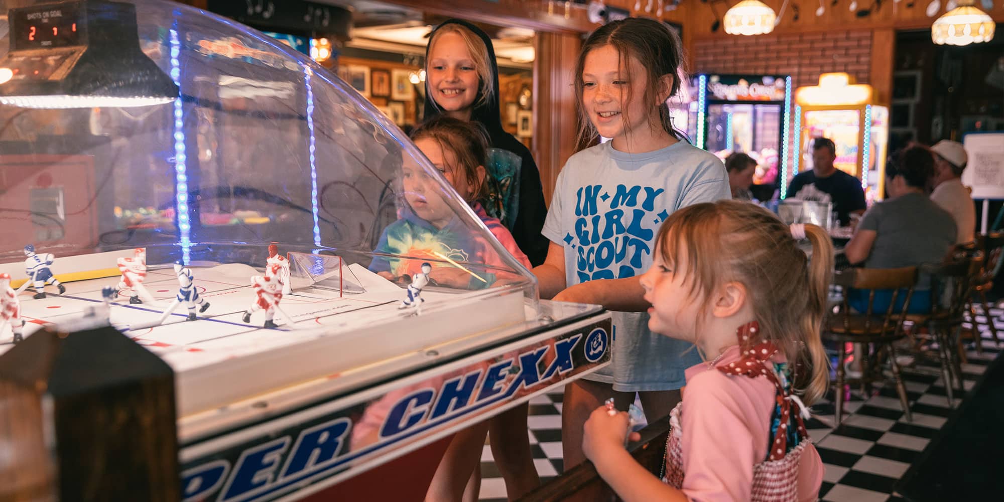 Children gathered around a Super Chexx hockey arcade game with smiles, in a lively setting with checkered flooring and neon lights.