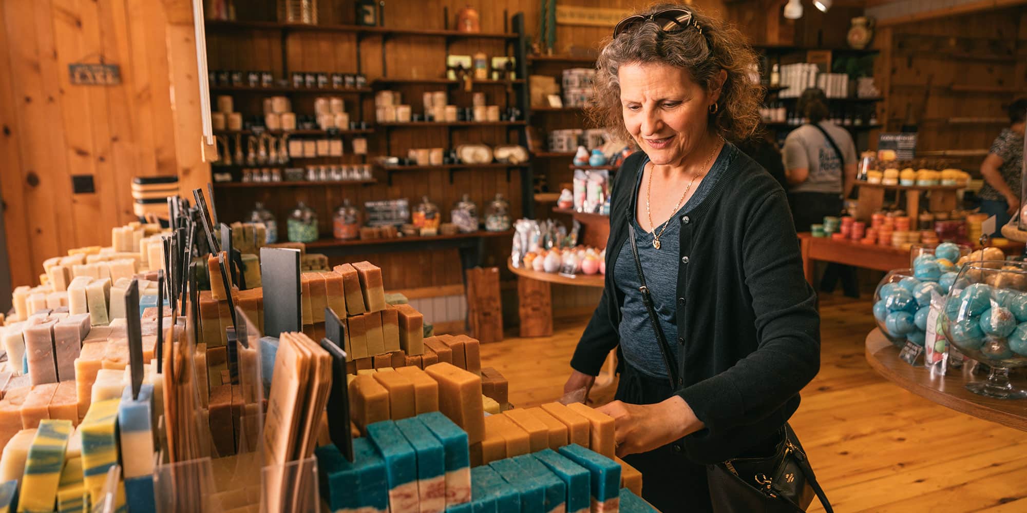 A woman browses and selects handmade soap bars in a rustic store filled with various soap and bath products.