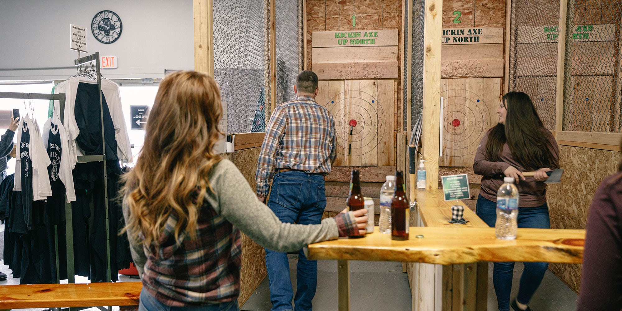 People at an axe-throwing venue, with two individuals aiming at targets and others watching while holding drinks.