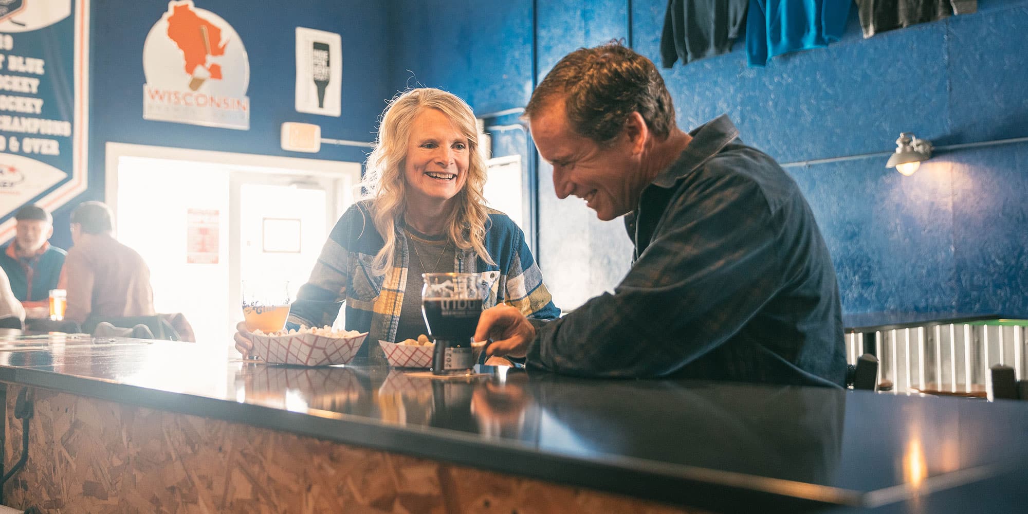 Two people sitting at a bar, laughing and talking. They have drinks and baskets of snacks in front of them, with a blue wall and Wisconsin-themed decor in the background.