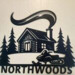 Silhouette of a cabin with smoke, surrounded by trees, and a snowmobile in front. Text reads "NORTHWOODS.