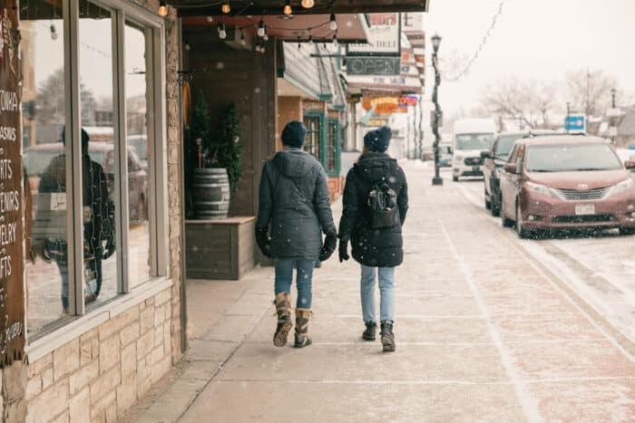 Two people wearing winter coats and backpacks walk down a snowy sidewalk in a small town. A few cars are parked on the street. Snowflakes are falling.