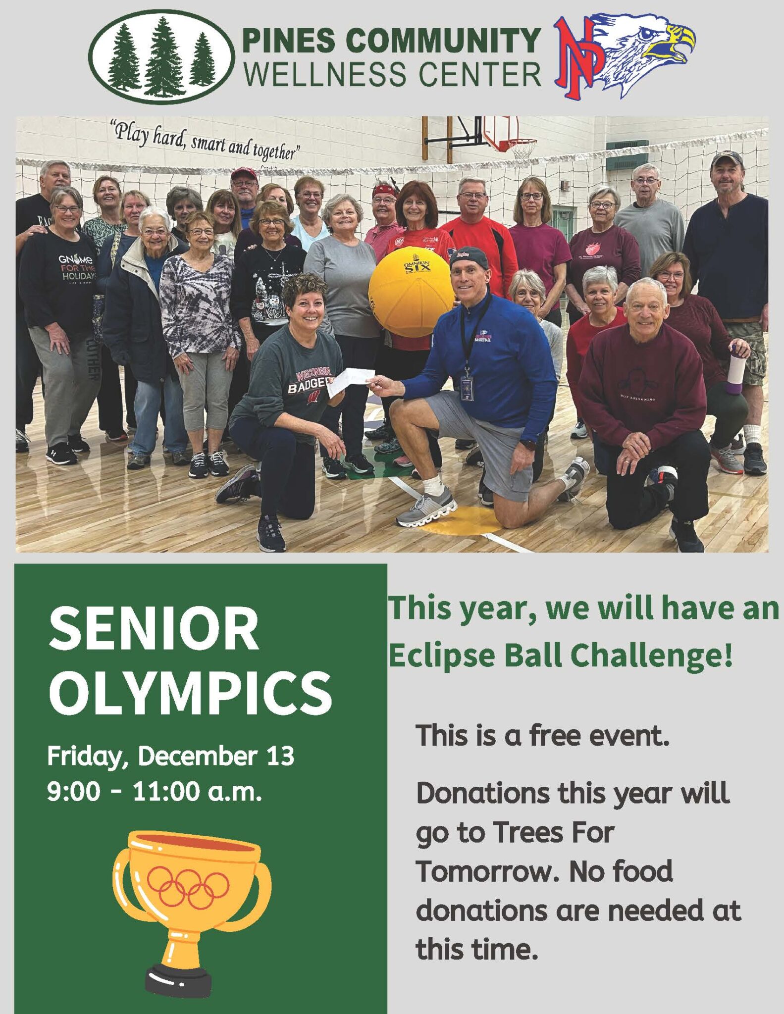 Group of people posing with a large yellow ball in a gymnasium, promoting the Senior Olympics event on Friday, December 13, from 9:00 to 11:00 a.m. at Pines Community Wellness Center.