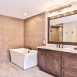 A bathroom with a large rectangular bathtub, double sink vanity with dark wood cabinets, a large mirror, and patterned wall tiles.