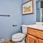 Bathroom with a toilet, wooden vanity, and sink. Blue walls feature a painting above the toilet. A towel bar is mounted on the wall.