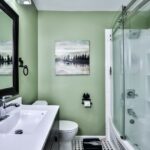 A bathroom with a green wall, a white vanity sink, a toilet, and a glass shower enclosure. A framed landscape painting hangs on the wall.