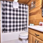 A rustic bathroom with wooden walls, a checkered shower curtain, white toilet, and a marble countertop with a sink. A mirror and decor are on the walls.