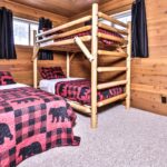 A cabin bedroom with wooden walls features a twin bed and a bunk bed, both with red and black bear-patterned quilts. Black curtains hang over small windows.