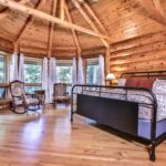 Wooden cabin bedroom with log walls, a metal bed, two chairs, a small table, and windows with white curtains. The ceiling is made of wooden beams. A lamp is on a dresser next to the bed.