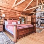 A cozy log cabin bedroom with a large wooden bed, stone fireplace, wooden ceiling beams, and a window with a view of trees.