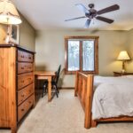 A rustic bedroom with a wooden bed, dresser, and desk. Two lamps are on either side, and a ceiling fan is above. A window shows a view of trees.