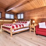 A cozy cabin bedroom with wooden walls and ceiling, featuring a bed with a red and white checkered duvet, a red sofa, a wooden dresser, and two nightstands with lamps.