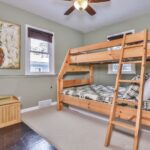 Bedroom with a wooden bunk bed, owl painting, and a small wooden box. Walls are green, and two windows are present. Ceiling fan above the room.