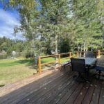 A wooden deck with a dining table and chairs, surrounded by tall trees, overlooks a grassy area and a lake in the background.