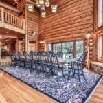 A spacious log cabin interior with a long dining table, surrounded by black chairs. A floral rug covers the wooden floor, and large windows offer a view of the greenery outside.