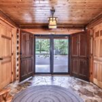 Wood-paneled entryway with double doors, stone flooring, a circular rug, wooden chair, and a glass door leading outside to a view of trees.