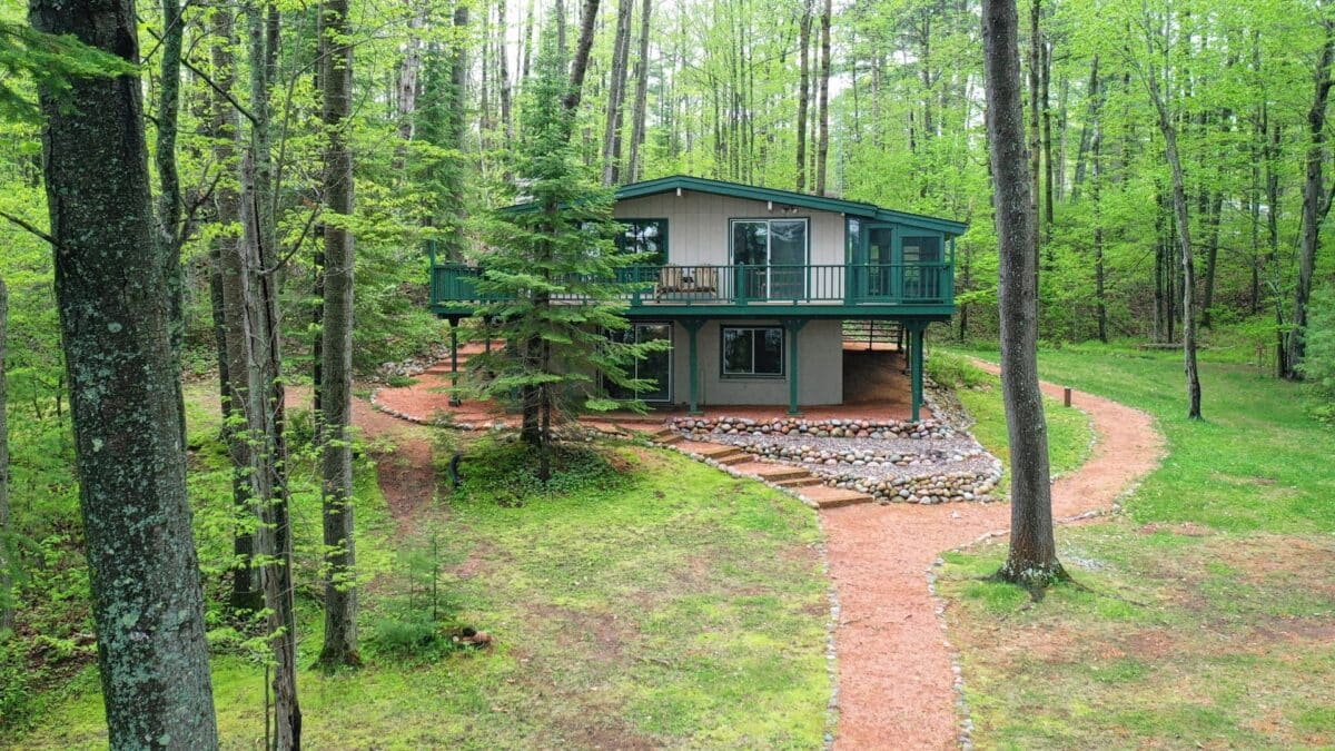 A two-story cabin surrounded by trees, with a stone path leading to the entrance, situated in a wooded area.