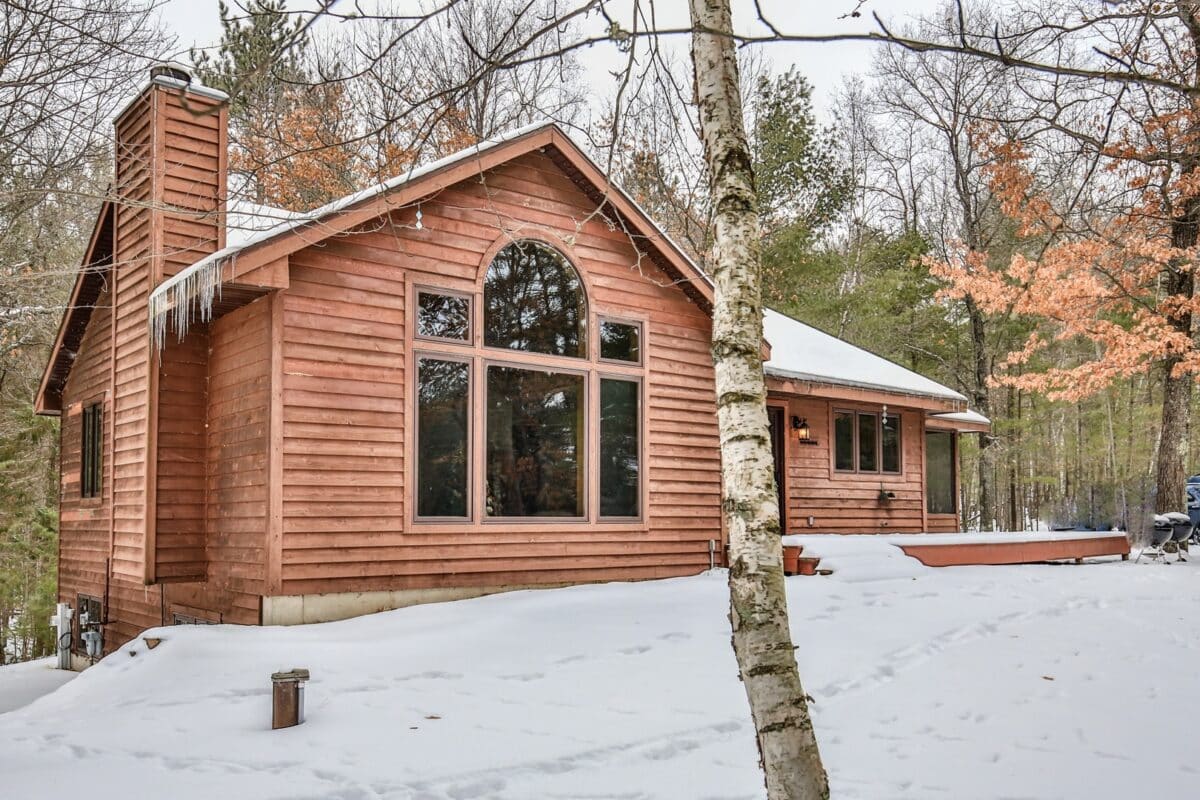 A wooden cabin with large windows is surrounded by snow and trees. A birch tree stands in the foreground.