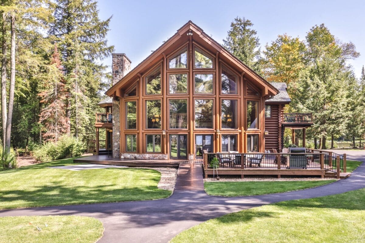 Large wooden cabin with floor-to-ceiling windows, set in a lush forest. A paved path leads to the entrance, surrounded by green grass and tall trees.