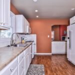 A kitchen with white cabinets, marble countertops, a white refrigerator, and an oven. The walls are a burnt orange color, and the floor is wood. A small patterned rug is on the floor.