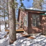 A brown cabin with large windows is surrounded by snow-covered ground and trees under a clear blue sky.