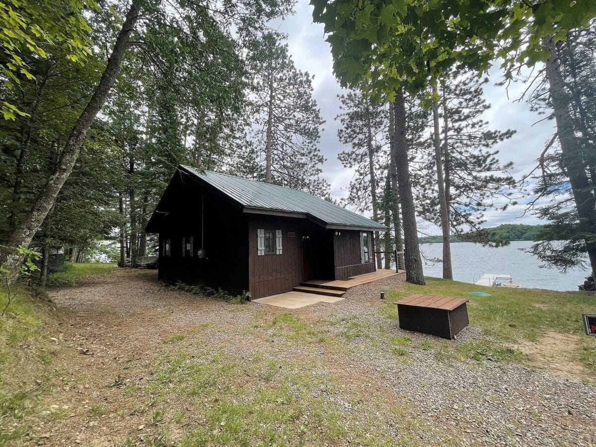 Small dark brown cabin with a green roof surrounded by trees near a lake. A gravel path leads to the cabin, and the lake is visible in the background under a cloudy sky.