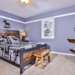 Bedroom with blue walls, featuring a bed with a plaid comforter, wooden bench, two lamps, a wall-mounted sink with a mirror, and a window with a snowy view.