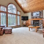 A cozy living room with large windows, leather furniture, a stone fireplace, and wooden accents, offering a view of the snowy forest outside.