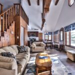 Living room with wooden beams, staircase, beige sofas, and a wooden coffee table. Dining area and kitchen visible in the background with large windows and blue-patterned curtains.