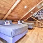 A rustic loft bedroom with two beds, wooden walls, and ceiling beams. A stone chimney is visible in the background, alongside a small seating area with a chair and table.