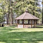 A wooden gazebo with a stone base stands in a grassy area surrounded by tall trees near a lake. Four black lounge chairs are set up on the lawn.