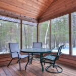 Screened porch with a round table and three chairs on a wooden floor, overlooking a snowy forest.