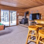 A cozy wooden cabin interior with a pool table, high table with stools, a wood-burning stove, and a large TV. Snow is visible outside through sliding glass doors.