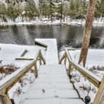 Snow-covered wooden steps lead to a frozen lake with a dock. Surrounding trees coated in snow line the shore.