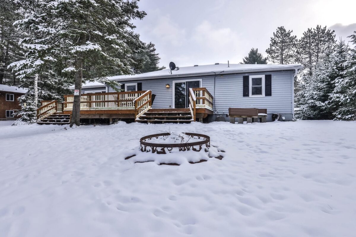 A single-story house with a wooden deck surrounded by snow, featuring a fire pit in the foreground and trees in the background.