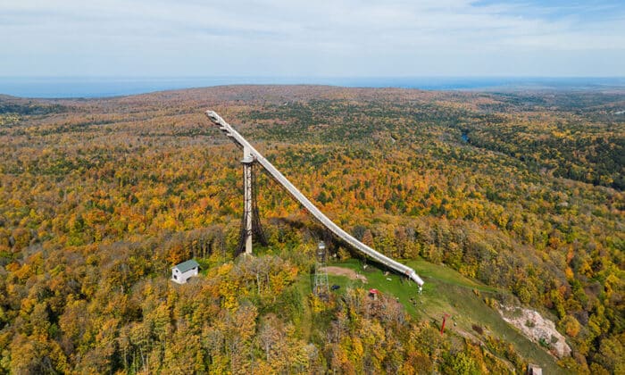 Aerial view of a large ski jump structure surrounded by a vast autumn forest landscape.