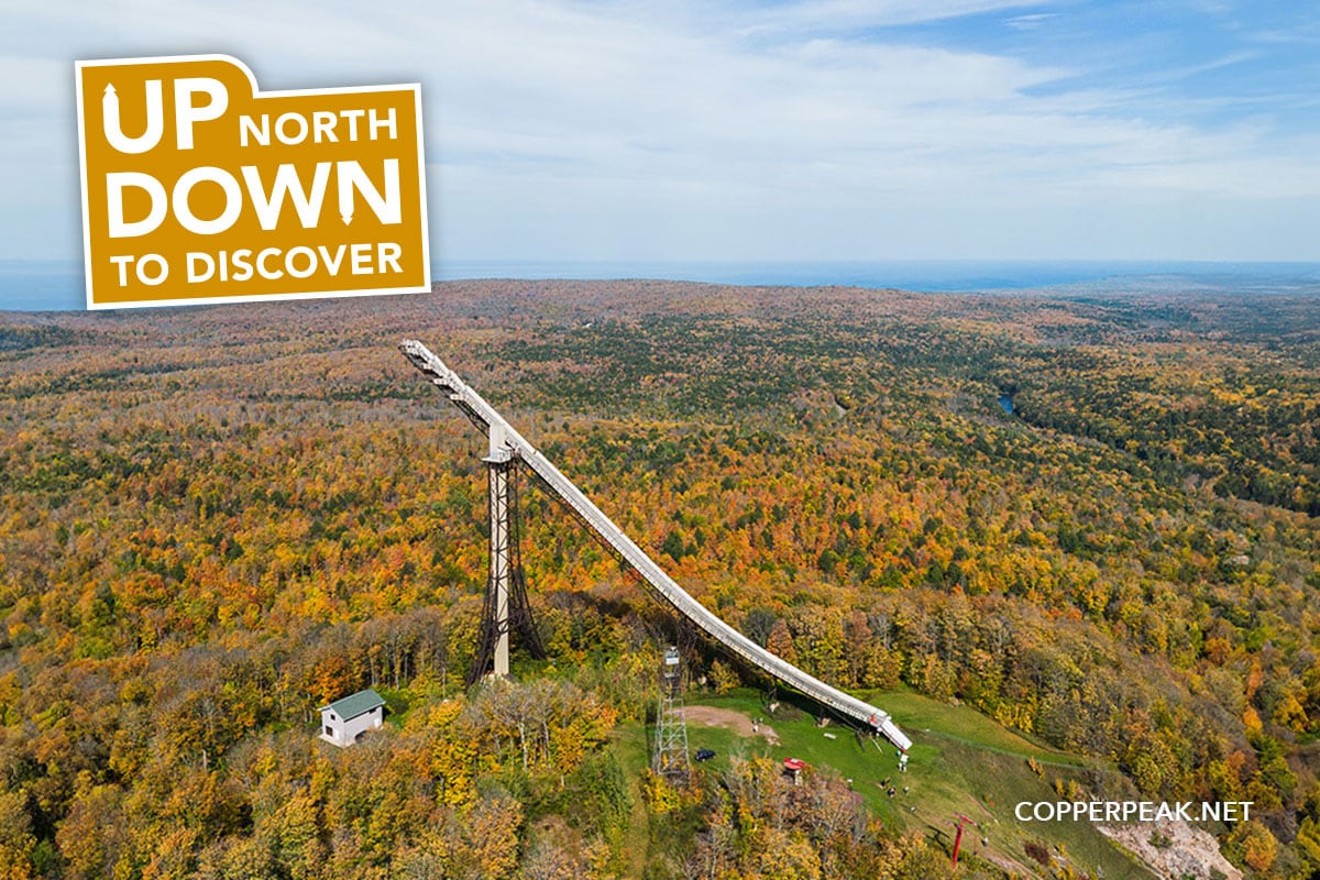 Aerial view of a ski jump surrounded by dense autumn forest. A sign reads "Up North, Down to Discover." Forest stretches into the distance.