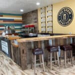 A rustic bar with a wooden counter, bar stools, a wall of beer taps, and a menu on the wall. A popcorn machine is near the bar. The sign reads "The Brew Station.