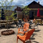 Outdoor seating area with orange and black chairs, picnic tables, and umbrella shades. Gravel ground and small green trees in the background under a partly cloudy sky.