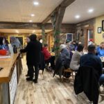 People dining in a cozy restaurant with wooden decor, seated at tables and a bar. Some are engaged in conversation, while others are eating or looking around.