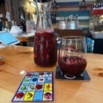 A carafe and glass of red sangria sit on a wooden bar. A colorful card and coaster are nearby. The background has a striped wall and various bar items.