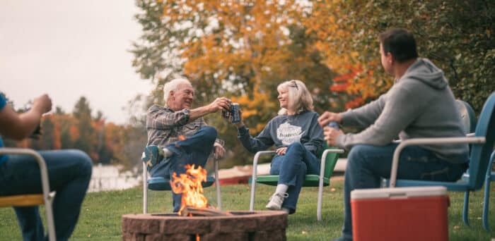 Four people sit around a fire pit outdoors on a grassy lawn, holding drinks and chatting, with autumn trees and a body of water in the background.
